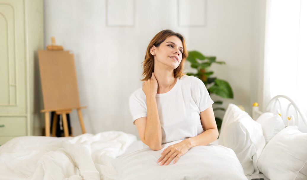 Woman waking up with neck pain, touching her shoulder while sitting on bed in morning light. Concept of bad sleeping posture, neck stiffness, discomfort and health or mattress related issues.